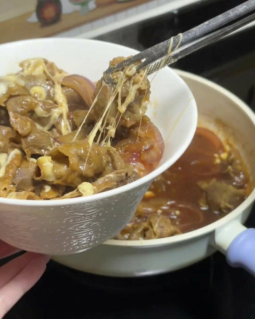 Szczypce podnoszące porcję ciągnącej się wołowiny teriyaki z serem z patelni i układające ją na misce z białym ryżem.