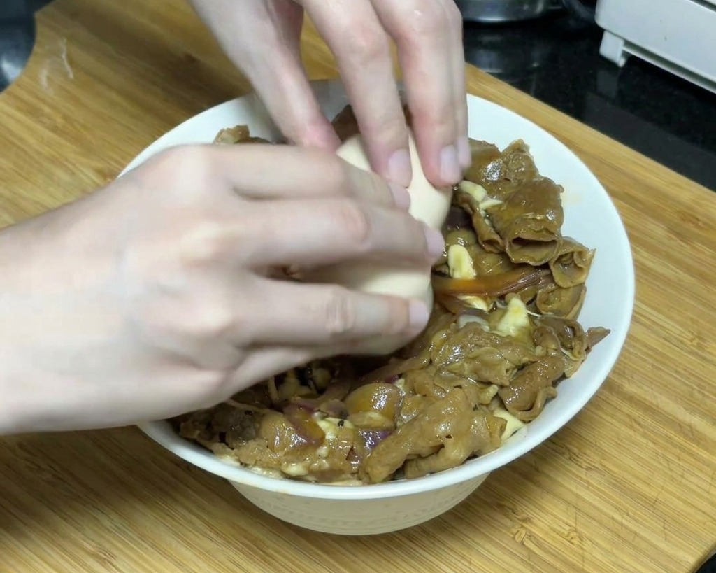 Dwie dłonie ostrożnie wbijające miękkie jajko onsen na środek miski z wołowiną teriyaki pokrytą roztopionym serem.