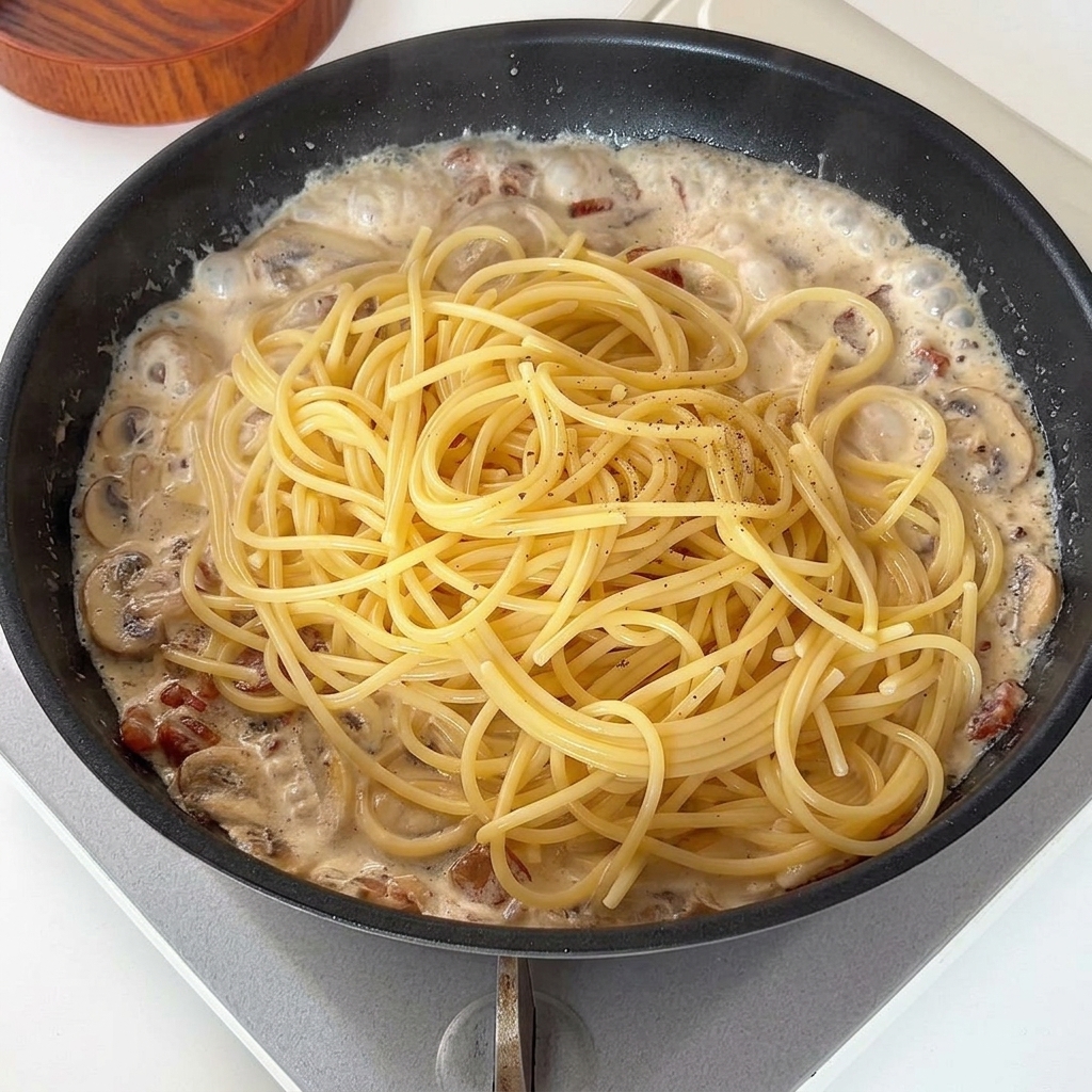 Spaghetti dodawane do kremowego sosu na patelni, posypane czarnym pieprzem.