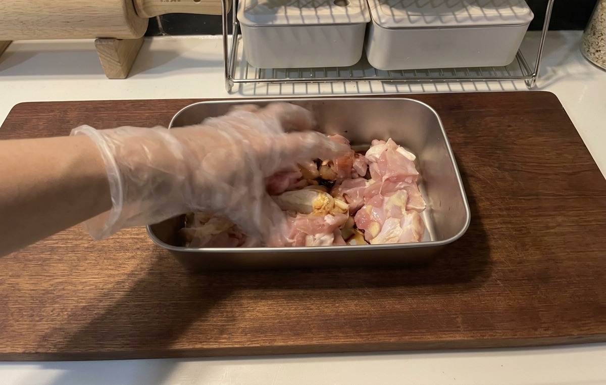 Dłoń w przezroczystej plastikowej rękawiczce mieszająca surowe kawałki kurczaka z ciemną marynatą z sosu sojowego na metalowej tacy leżącej na drewnianej desce.