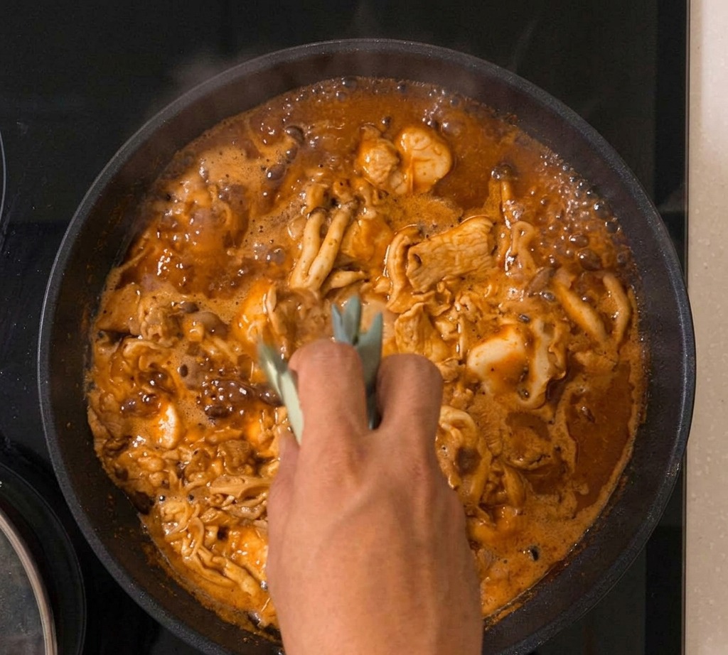 Mieszanka oyakodon bulgocze na patelni z widoczną zieloną cebulką pośród ścinających się jajek i kurczaka.