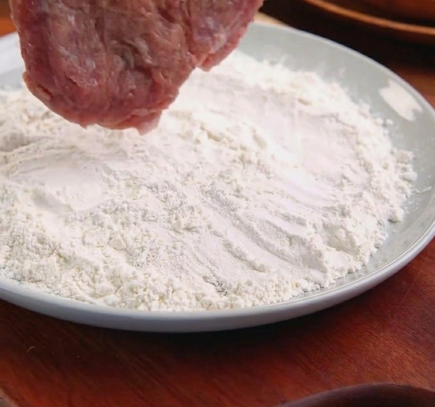 Kotlet wieprzowy trzymany szczypcami, zanurzany w talerzu z mąką.