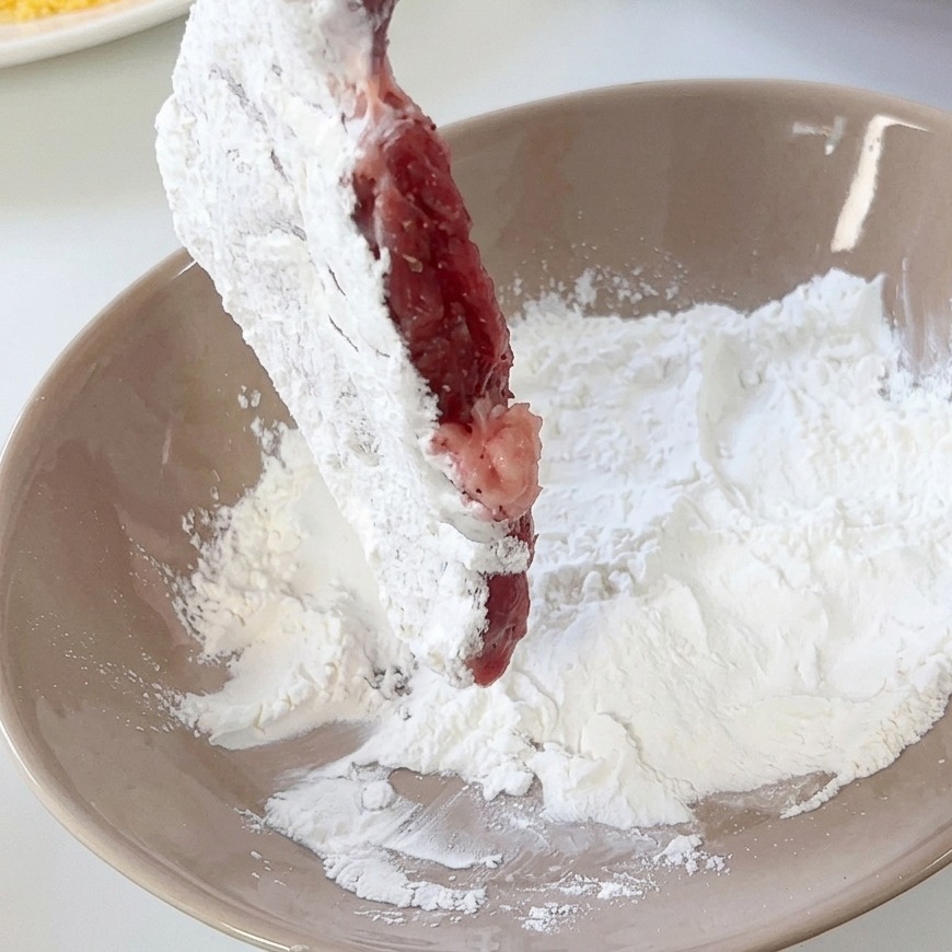 Kotlet wieprzowy obtaczany w białej mące w jasnobrązowej misce.
