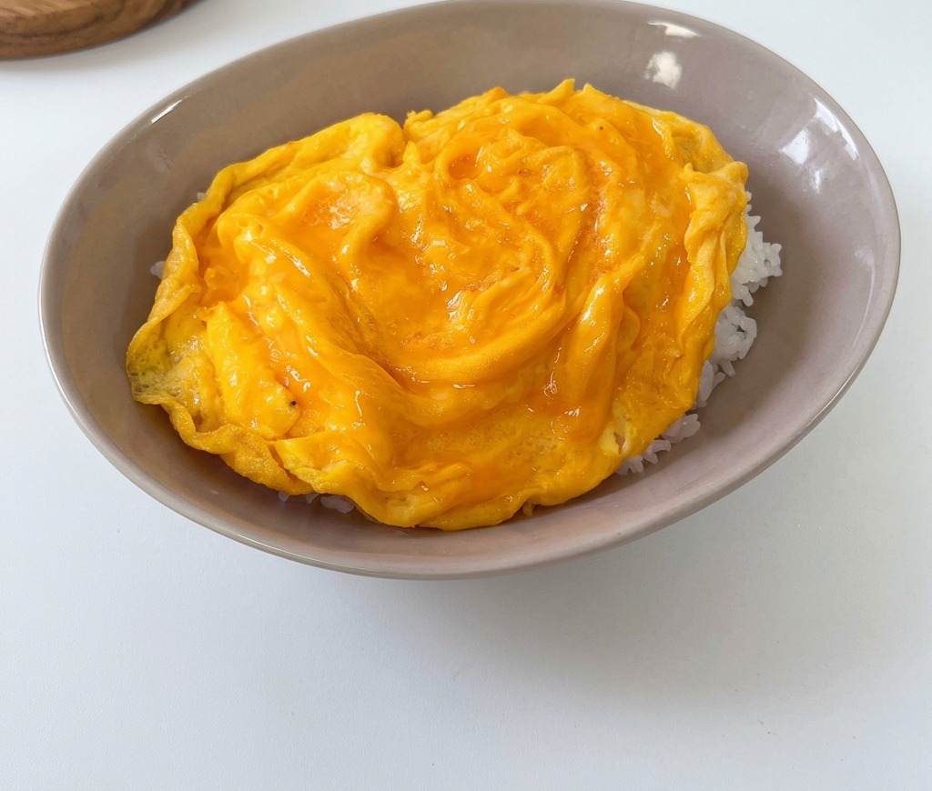 Puszysty, miękki żółty omlet ułożony na porcji białego ryżu w płytkiej, szarej misce.