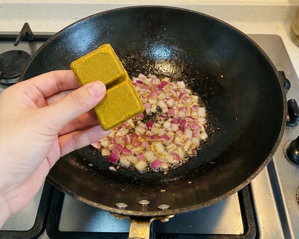 Osoba trzymająca dwie brązowe kostki curry nad wokiem z podsmażoną czerwoną cebulą.