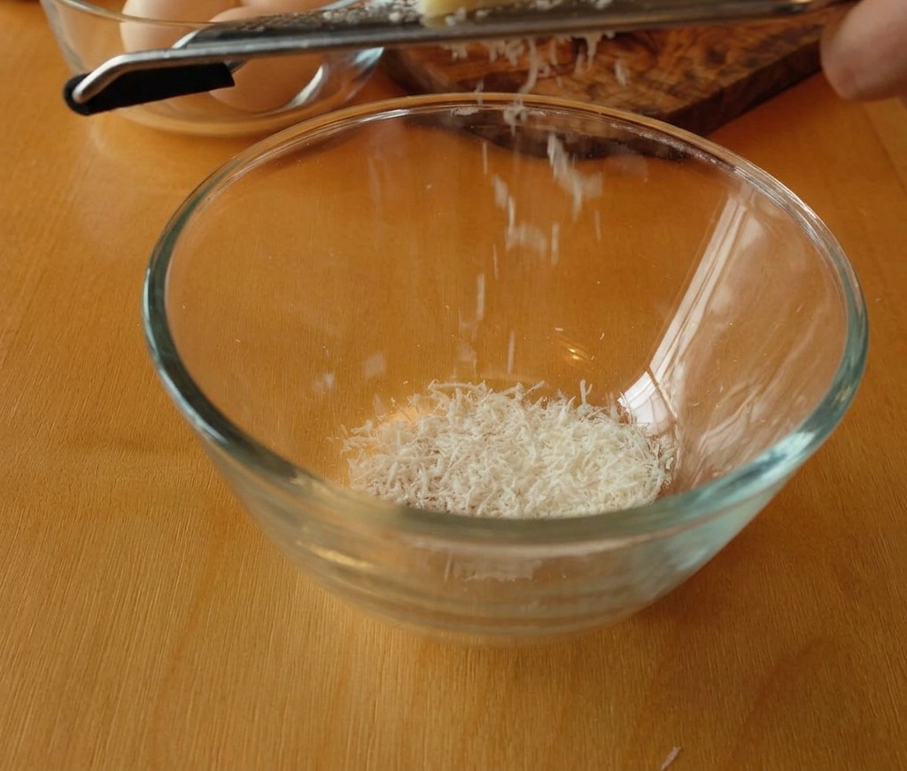 Blok sera Pecorino Romano ścierany do szklanej miski, tworzący puszyste, białe wiórki.