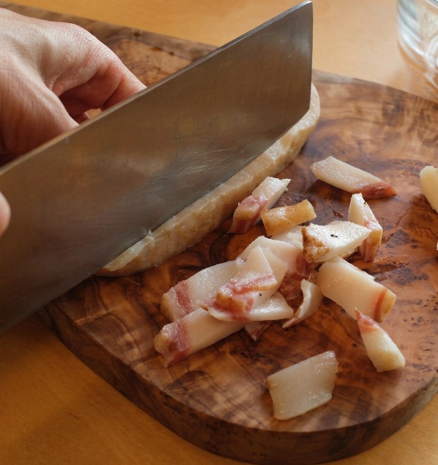 Ostry nóż odcinający grubą, ciemną skórę z kawałka guanciale na drewnianej desce.