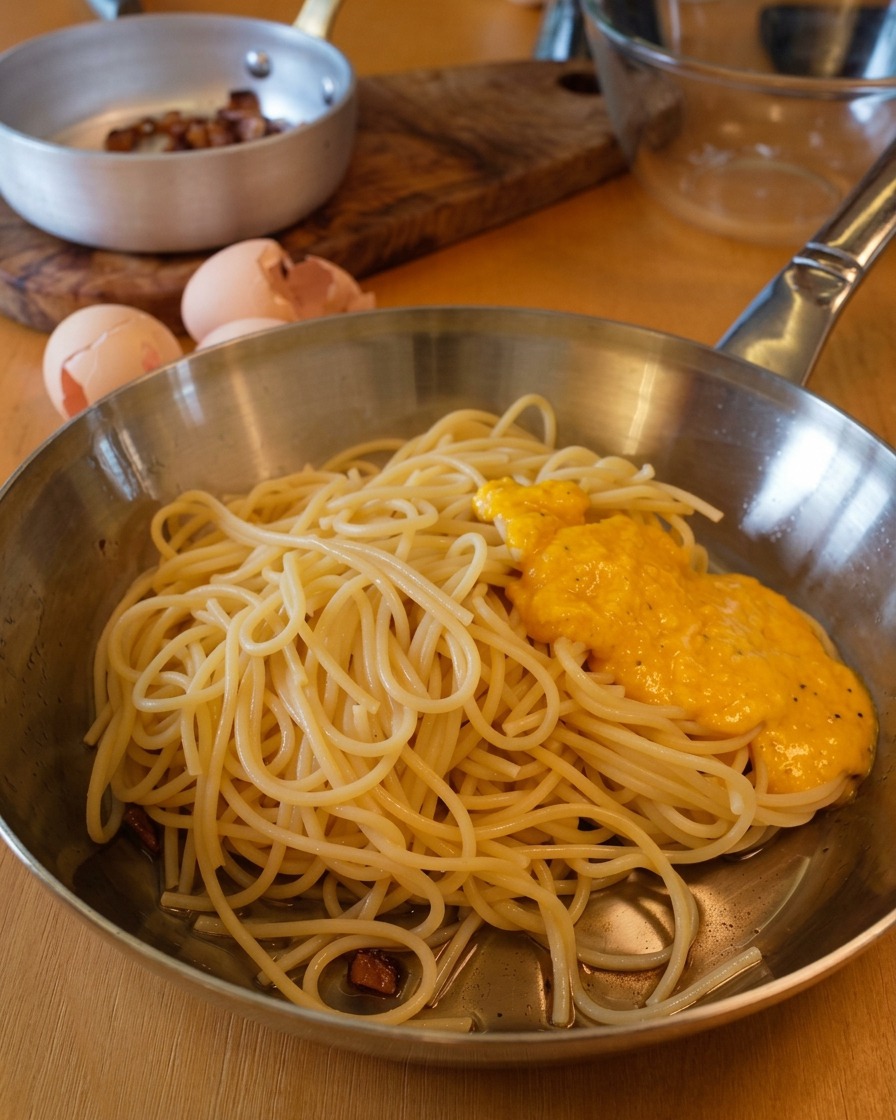 Gęsty, złocisty sos z jajek i sera wlany na porcję spaghetti na metalowej patelni.