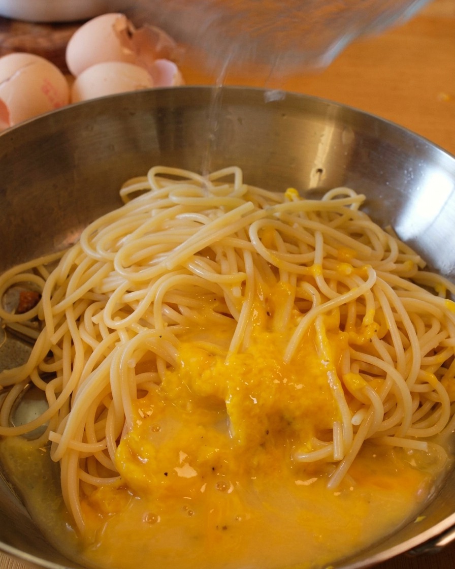 Spaghetti na patelni pokrywane żółtym sosem podczas dolewania wody z gotowania makaronu.