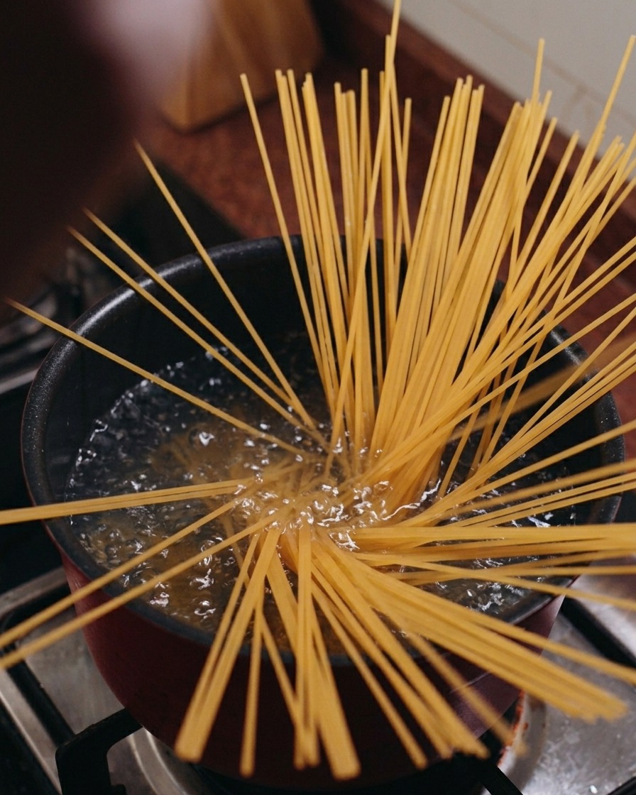 Suchy makaron spaghetti ustawiony pionowo w garnku z wrzącą wodą, rozchodzący się w kształt gwiazdy.