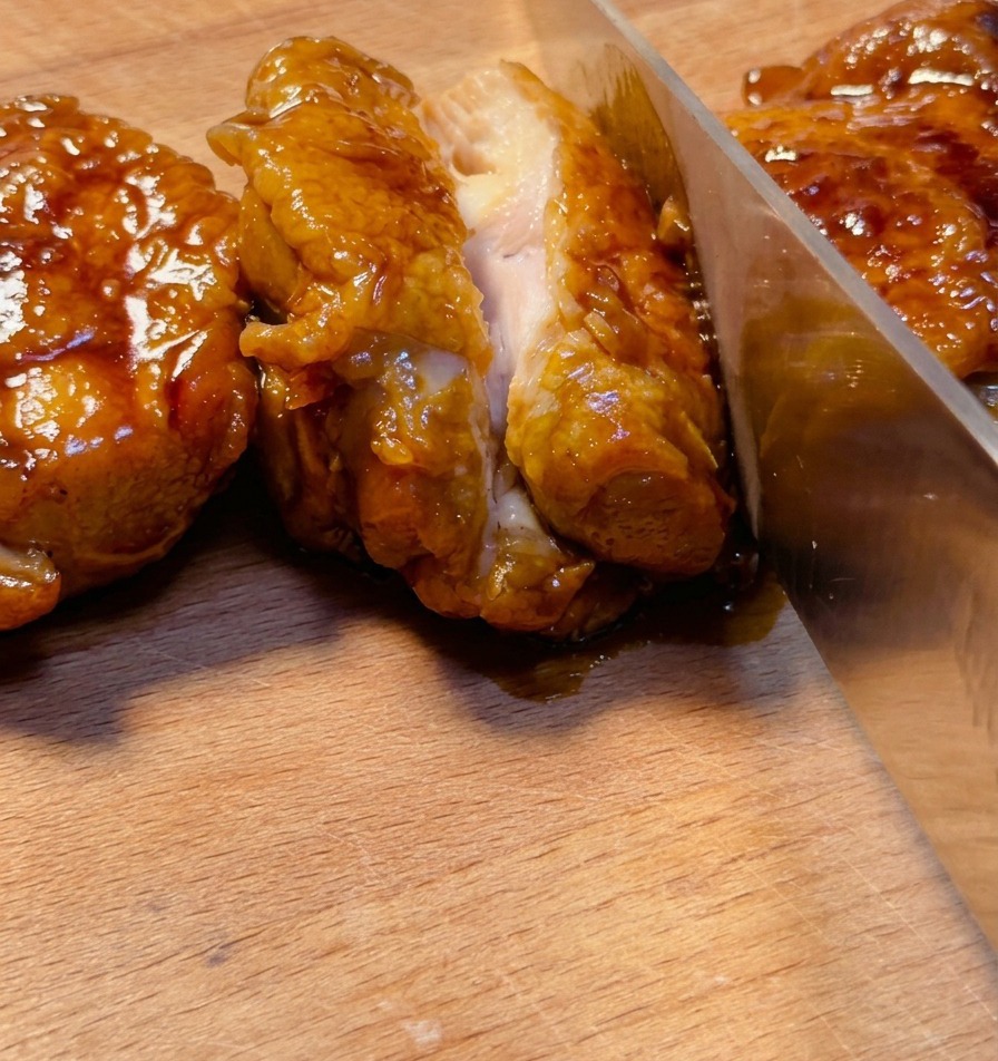 Ostry nóż krojący pokryte błyszczącą glazurą udko teriyaki na drewnianej desce, ukazując soczyste wnętrze.