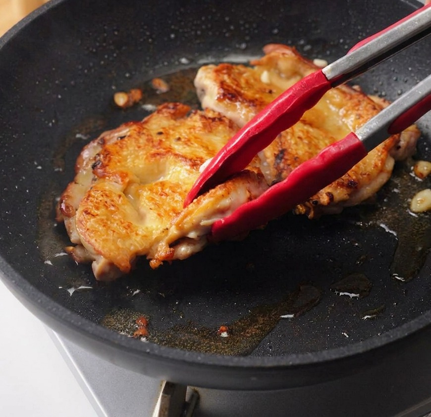 Dłoń wlewająca ciemny sos teriyaki ze szklanej miseczki bezpośrednio na usmażone udka z kurczaka na patelni.
