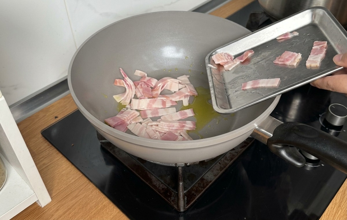 Surowe paski boczku wsypywane z metalowej tacki na szarą patelnię z oliwą.