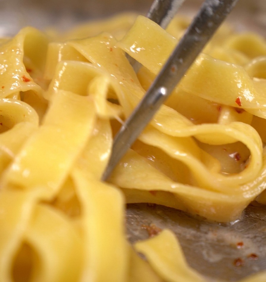 Makaron tagliatelle obracany metalowymi szczypcami na patelni z kawałkami boczku i delikatnie wrzącą wodą po makaronie.