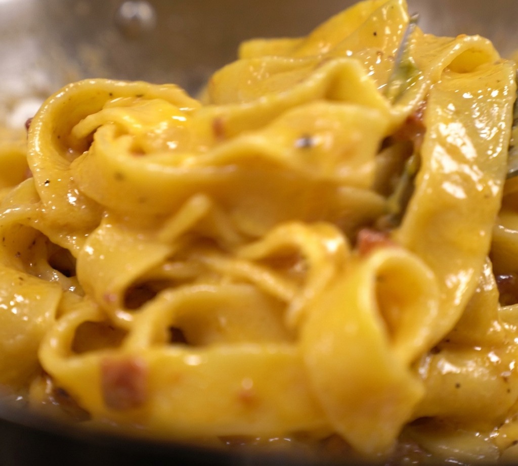 Makaron tagliatelle pokryty gęstym, jasnożółtym kremowym sosem jajeczno-serowym z widocznymi kawałkami boczku na patelni.