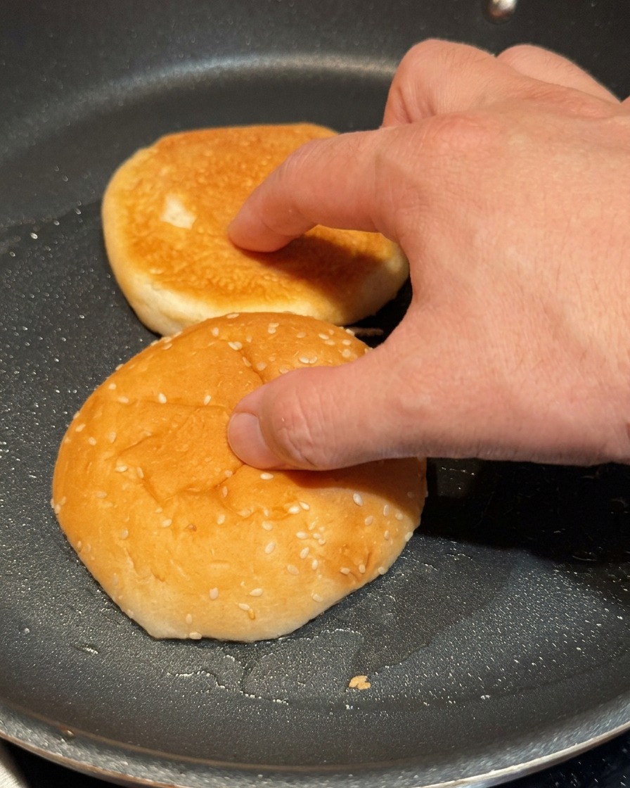 Dłoń lekko dociskająca dwie połówki bułek do burgera do gorącej czarnej patelni.