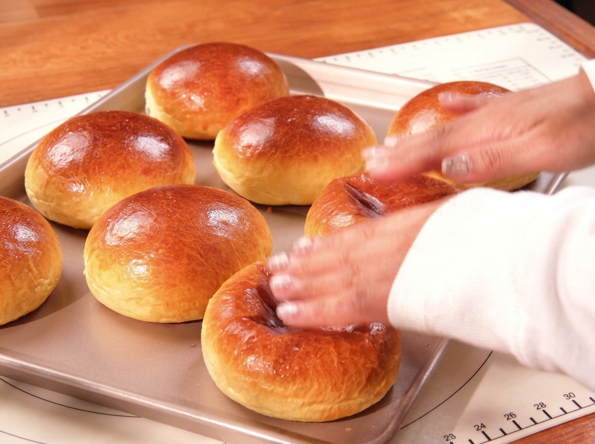 Dłoń dotykająca świeżo upieczonych, złocistych bułek brioche na blasze.