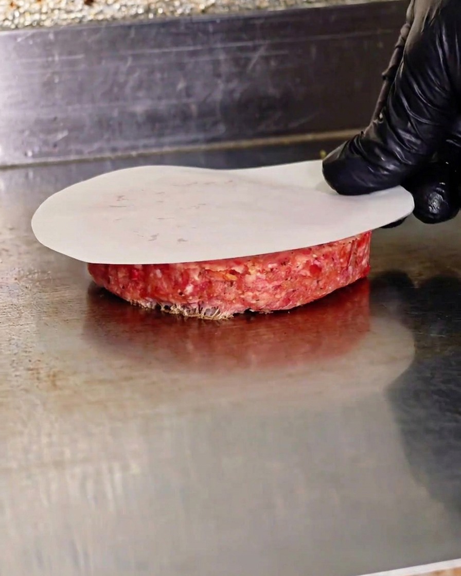 Ręka dociskająca surowy kotlet wołowy za pomocą prasy przez papier do pieczenia na gorącej płycie.