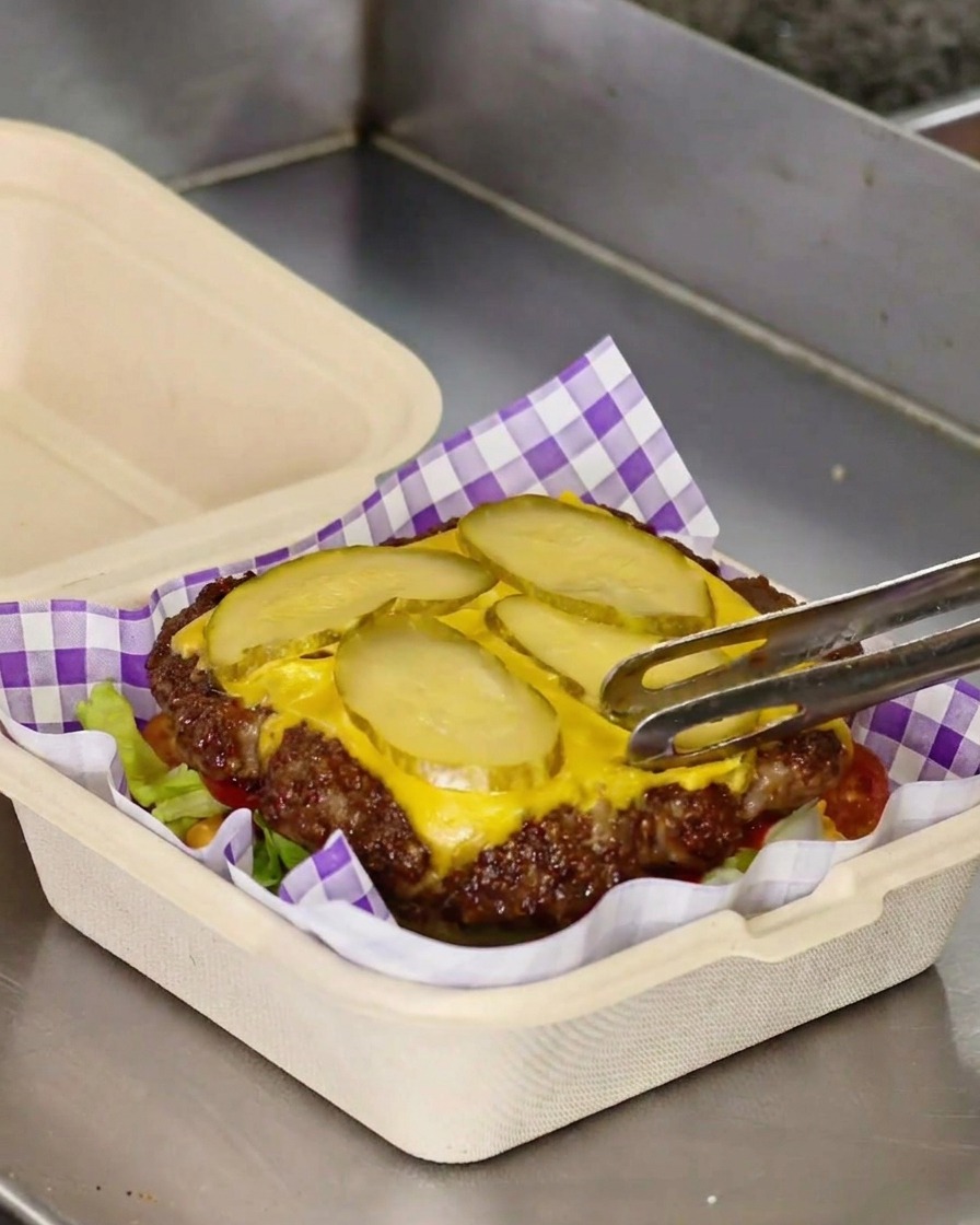 Szczypce przenoszące upieczony kotlet smash z serem i ogórkami na warstwę sałaty i pomidora.
