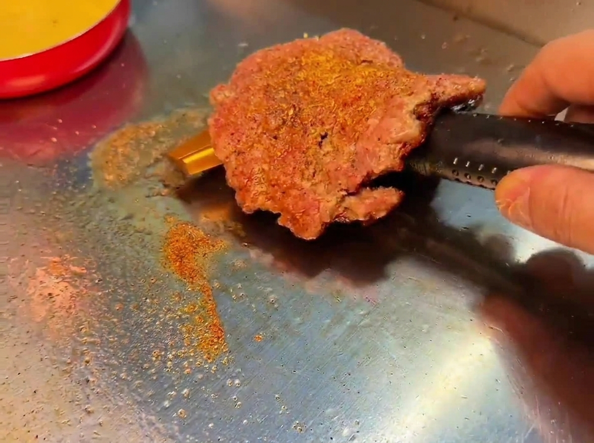 Smashed burger obrócony na grillu, ukazujący głęboko brązową, przypieczoną skórkę.