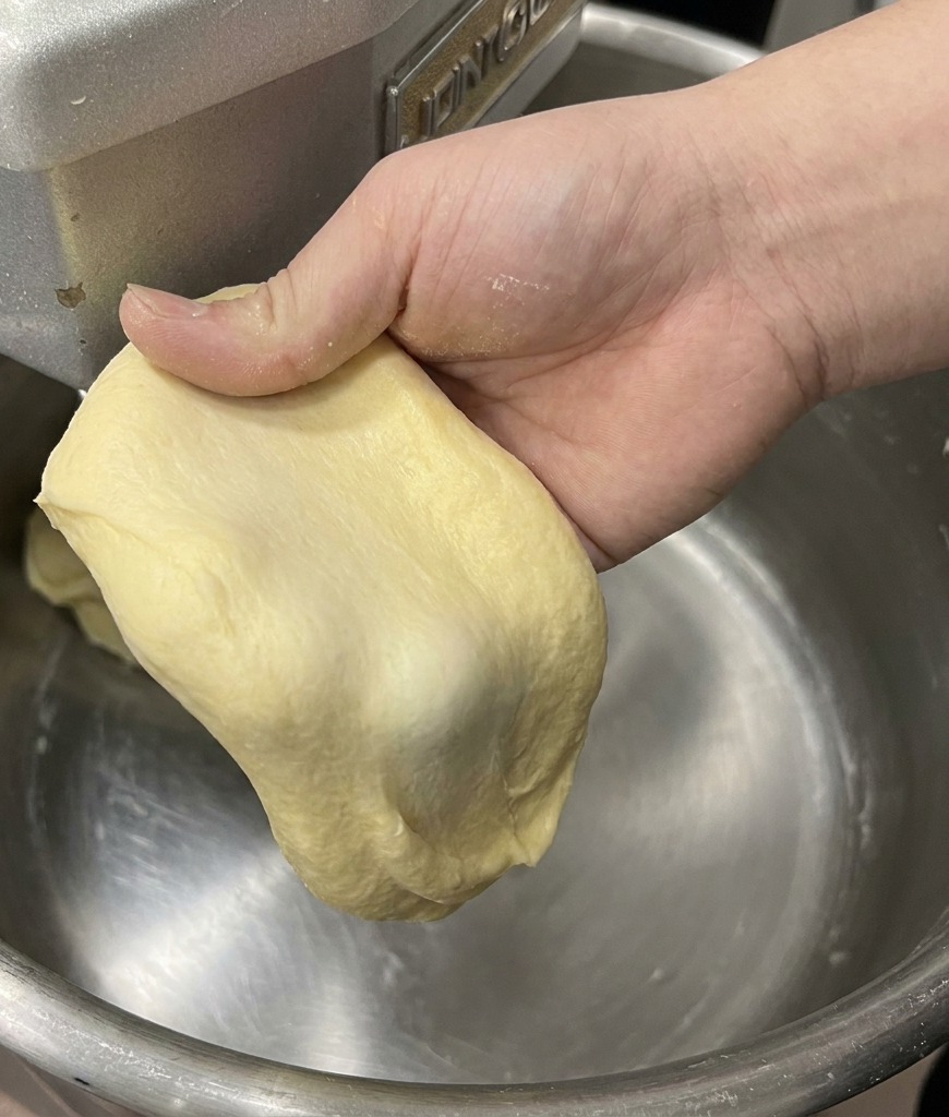 Ręka rozciągająca kawałek jasnego ciasta brioche, pokazująca cienką, mocną membranę (test okienka).