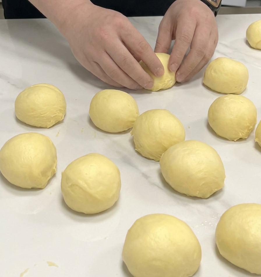 Dłonie formujące małe porcje żółtego ciasta brioche w kulki na białym marmurowym blacie.