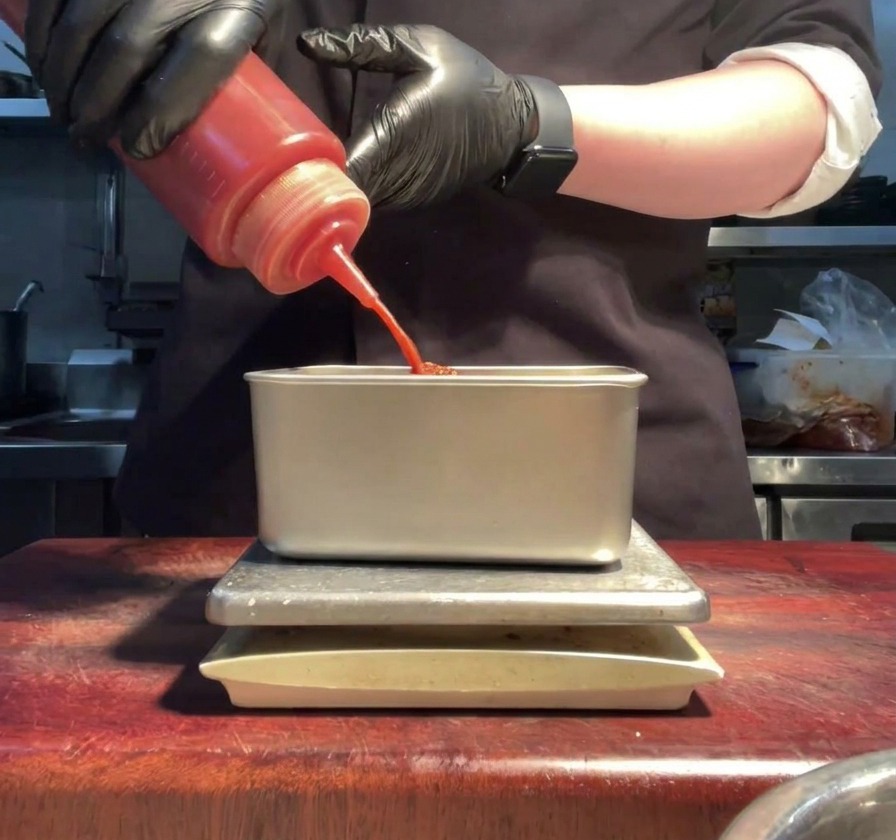 Ręce w rękawiczkach wyciskające jaskrawoczerwony sos sriracha do metalowego pojemnika.