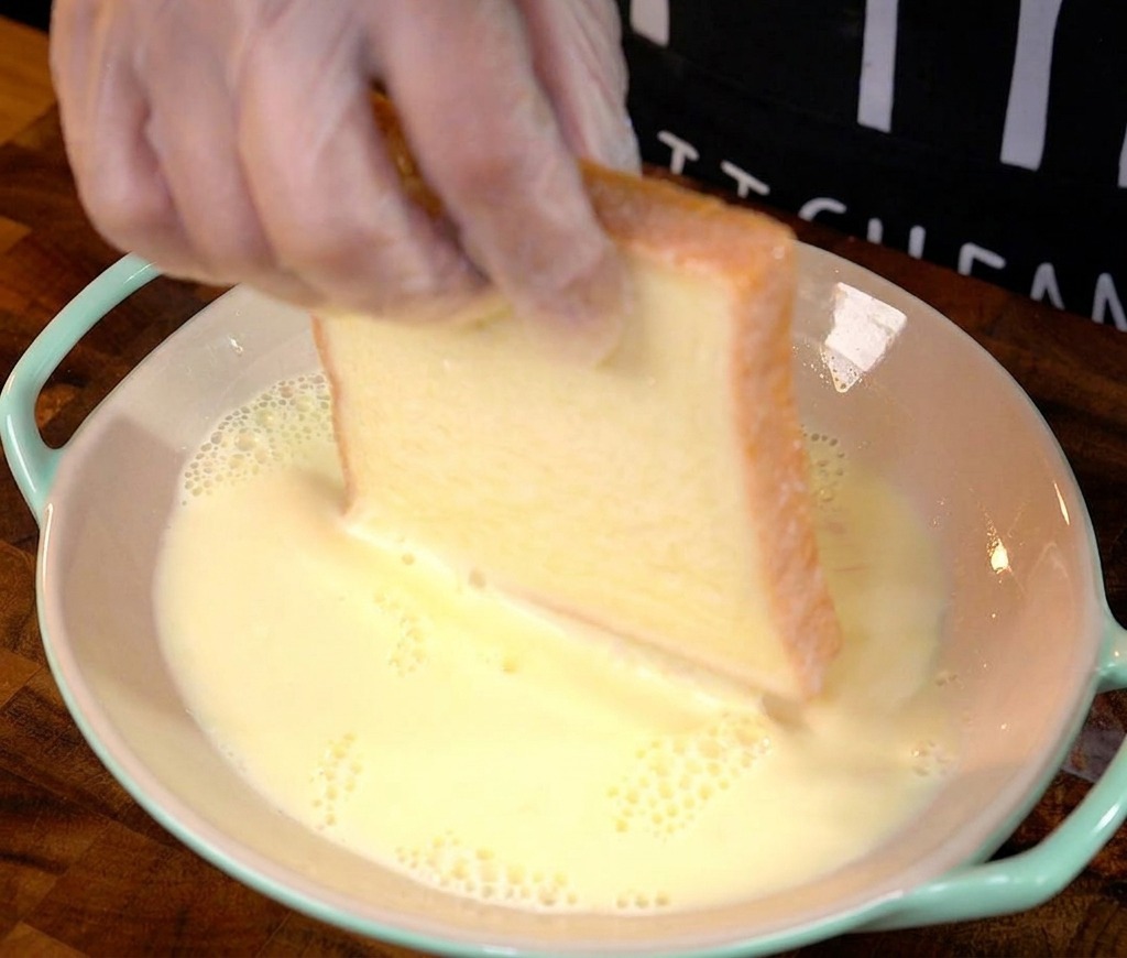 Ręka zanurzająca grubą kromkę białego chleba w bladożółtej masie jajecznej.