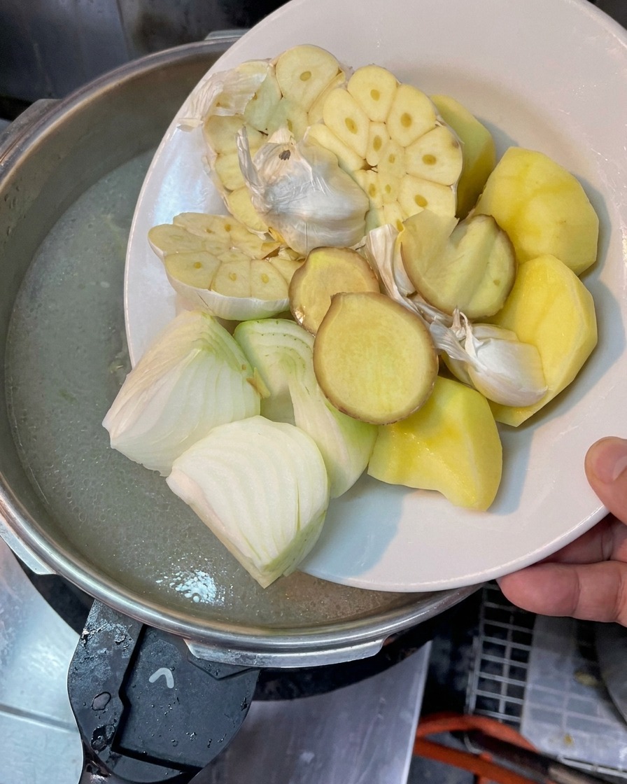 Talerz z przekrojonym czosnkiem, cebulą, plastrami imbiru i kawałkami ziemniaka trzymany nad garnkiem z gotującym się wywarem.