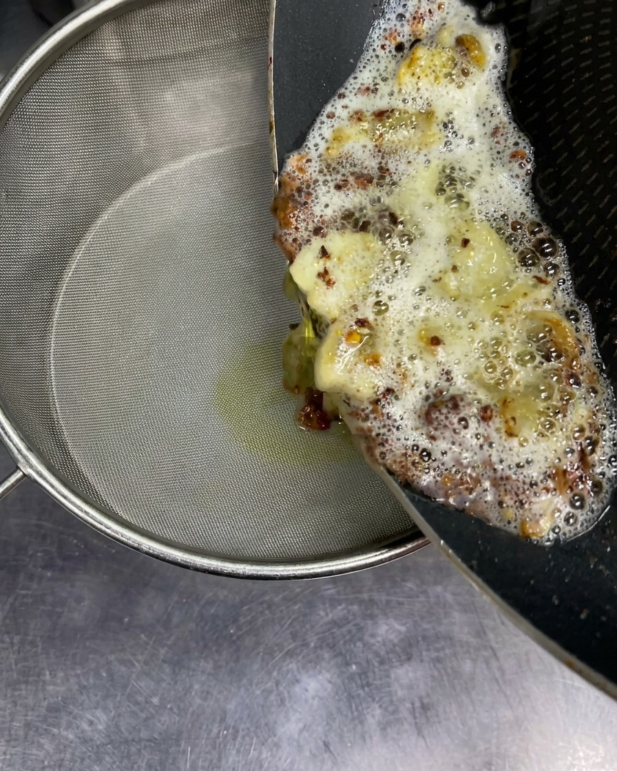 Gorący, bąbelkujący olej aromatyczny ze smażonym czosnkiem przelewany z patelni przez sitko.