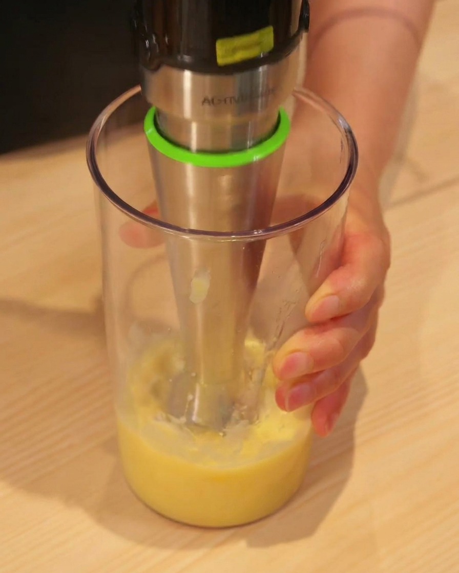 Blender ręczny miksujący kawałki żółtego ananasa na gładki sok w wysokim naczyniu.