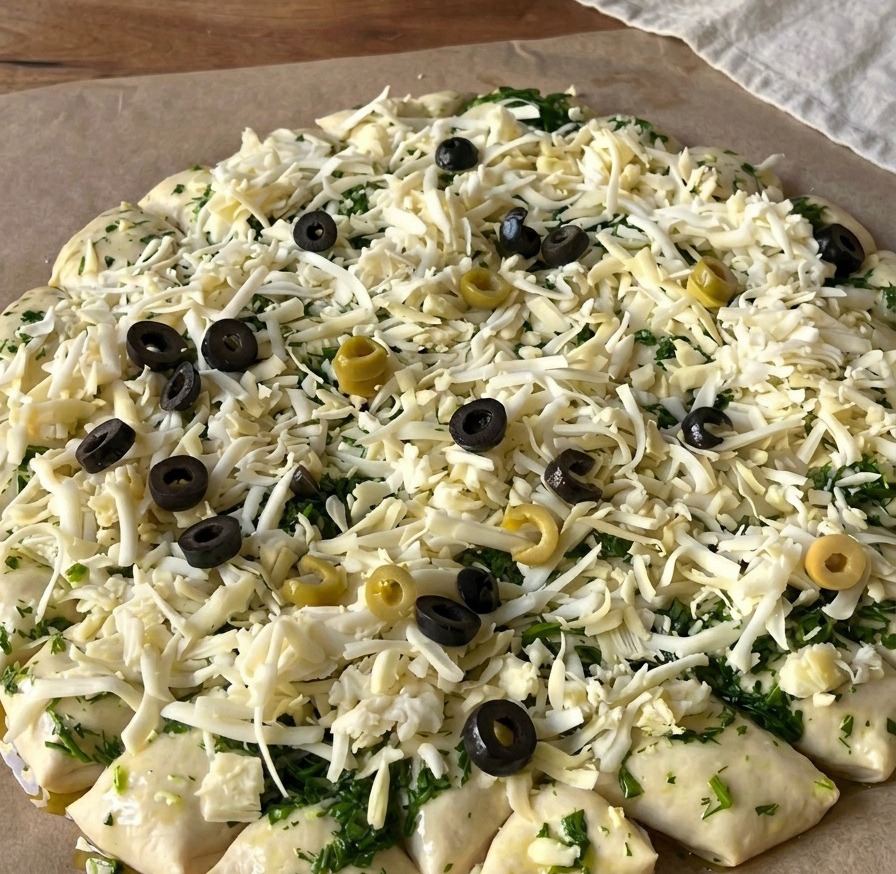 Widok z góry na surowe ciasto na pizzę pokryte tartym serem oraz licznymi plasterkami czarnych i zielonych oliwek.