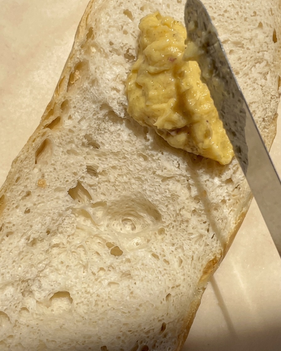 Zbliżenie na nóż rozsmarowujący grubą, kremową warstwę masła z pieczonym czosnkiem na połówce bagietki.