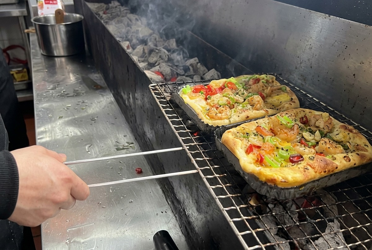 Prostokątna pizza z krewetkami i warzywami układana na długim ruszcie grilla węglowego w profesjonalnej kuchni.