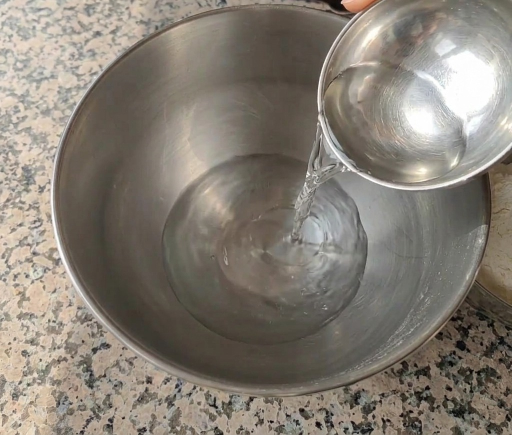 Woda nalewana z małej metalowej miski do dużej misy miksera ze stali nierdzewnej na granitowym blacie.
