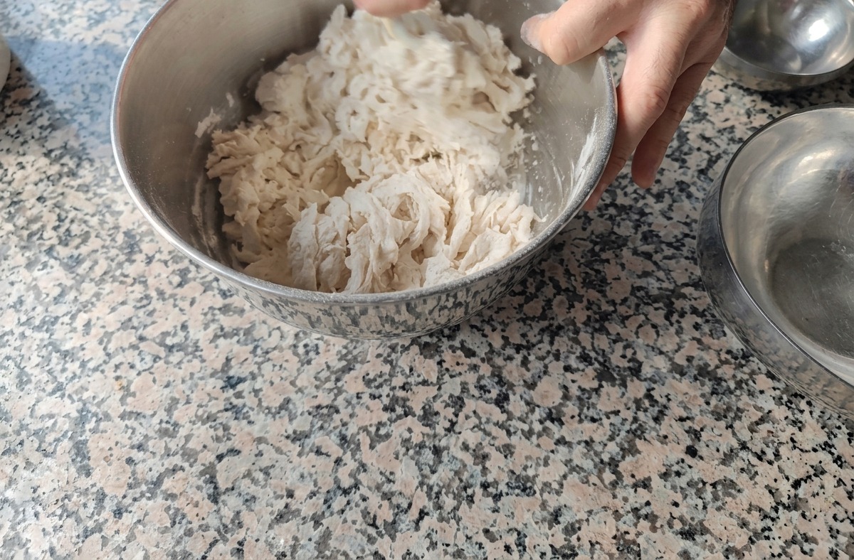 Dłonie mieszające mąkę z wodą w metalowej misie w celu uzyskania rwanego, nieregularnego ciasta Biga.