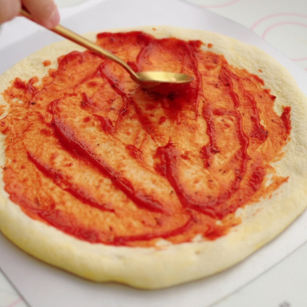 Złota łyżka rozprowadzająca jasnoczerwony sos pomidorowy kolistymi ruchami na surowym spodzie pizzy.