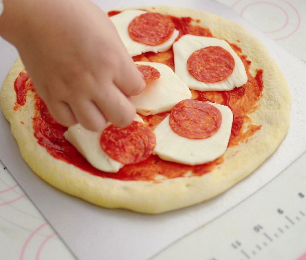 Dłoń układająca cienkie, okrągłe plasterki salami na spodzie pizzy z sosem pomidorowym i mozzarellą.