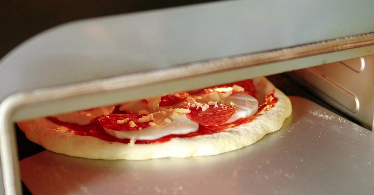 Surowa pizza z salami zsuwana ze srebrnej metalowej łopaty prosto do rozgrzanego wnętrza pieca do pizzy.