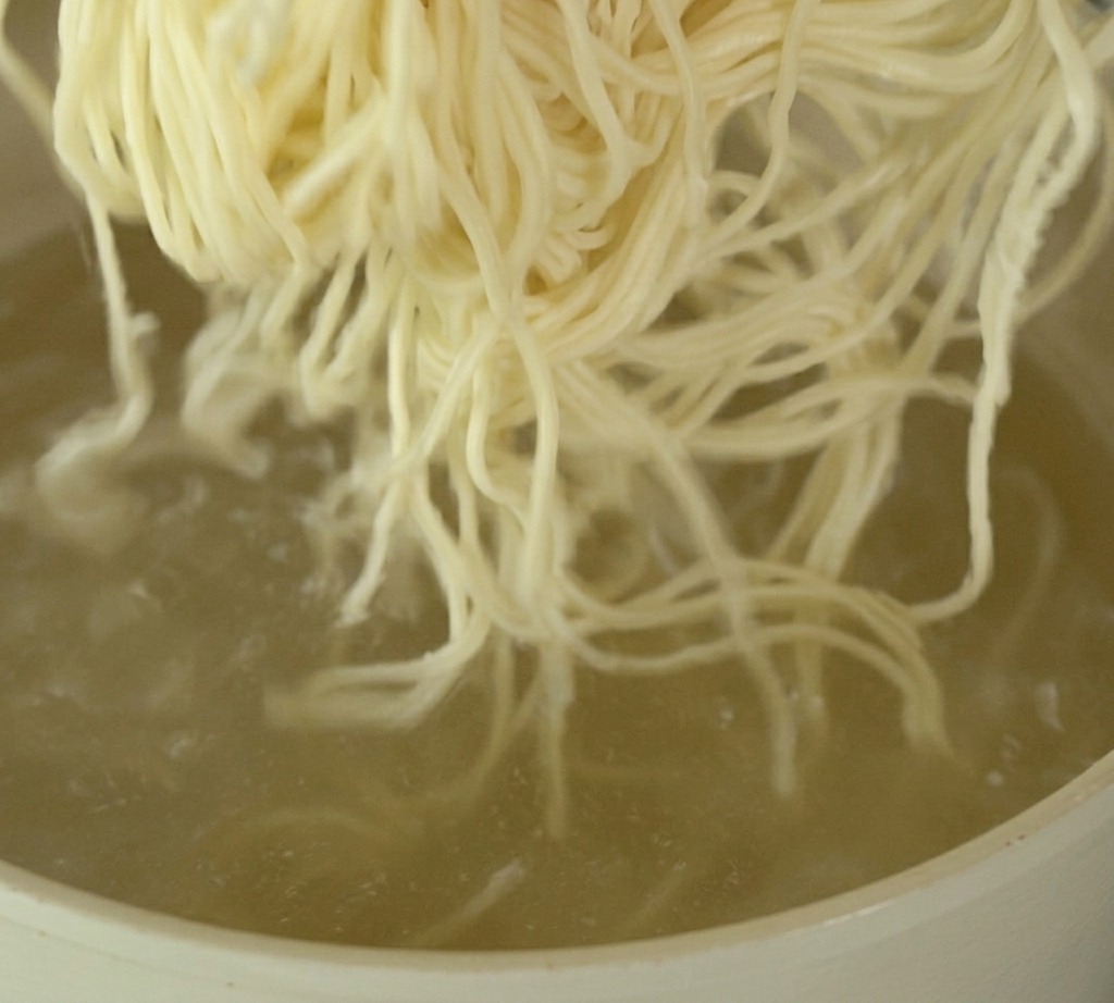 Porcja cienkiego, jasnego makaronu ramen zanurzana w garnku z intensywnie gotującą się wodą.