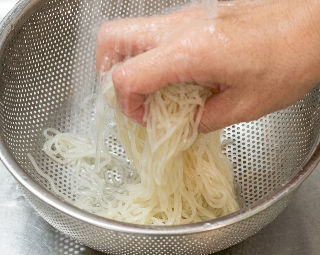 Dłoń płucząca jasny makaron ramen pod strumieniem zimnej wody w stalowym durszlaku.
