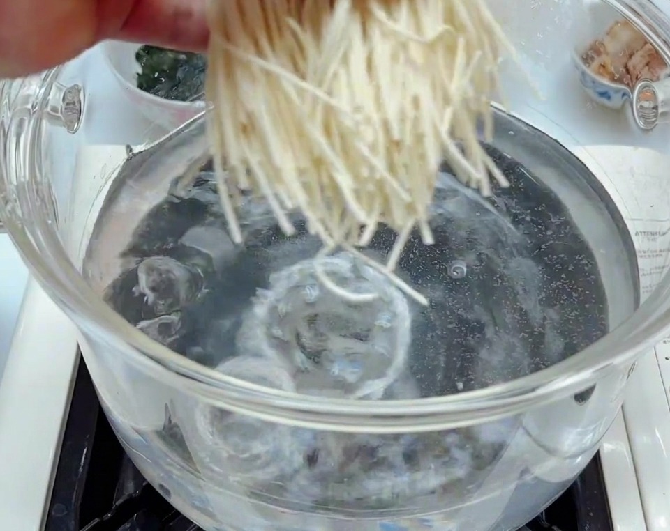 Świeży, jasny makaron ramen wrzucany do szklanego garnka z mocno wrzącą wodą.