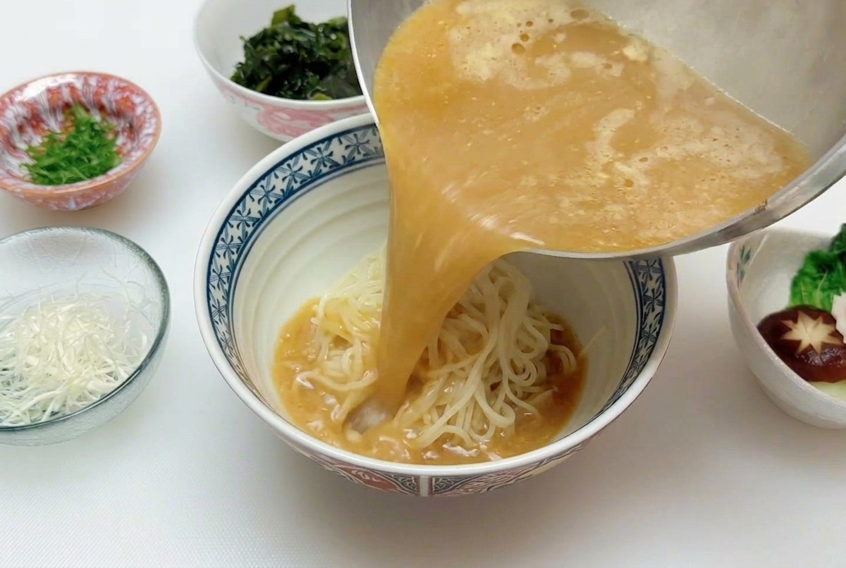 Strumień gorącego, złocistobrązowego bulionu miso wlewanego z rondelka bezpośrednio na ułożony w misce makaron ramen.