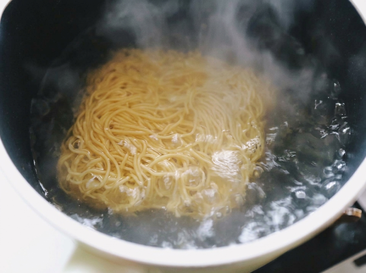 Falowany makaron ramen zanurzony w garnku z mocno gotującą się wodą, z której unosi się para.
