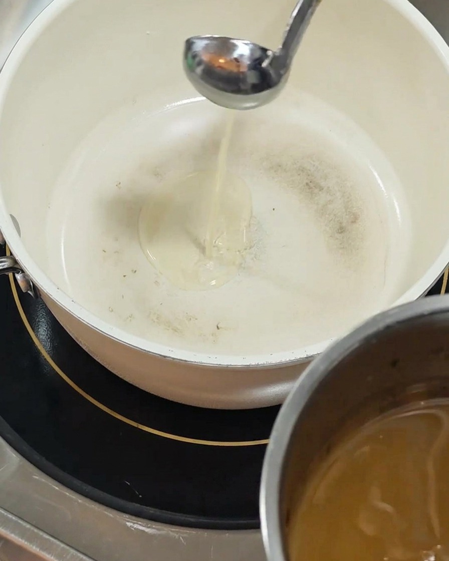 Srebrna chochla nalewa przejrzysty, żółty olej do białego garnka z powłoką ceramiczną, stojącego na czarnej kuchence indukcyjnej.
