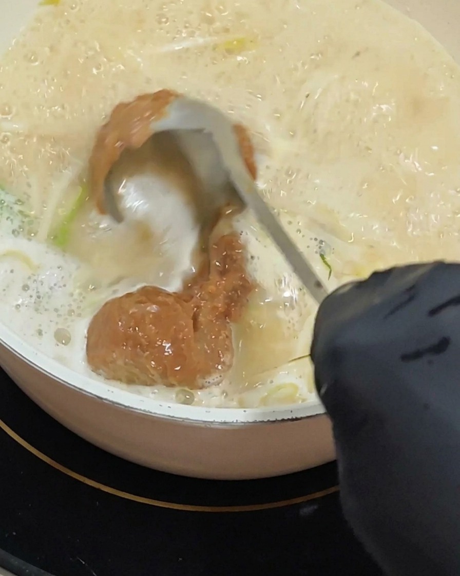 Dłoń w rękawiczce używająca chochli do wmieszania grubej warstwy czerwonobrązowej pasty miso w bulgoczącą, kremową bazę zupy.