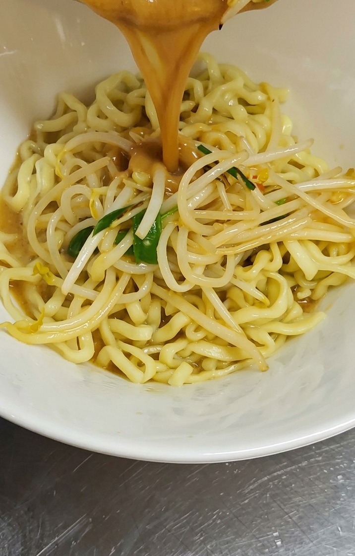 Strumień gęstego, brązowego bulionu miso i podsmażonych kiełków wylewany z patelni do białej miski wypełnionej pofalowanym, żółtym makaronem ramen.