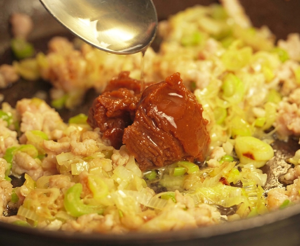 Srebrna łyżka nakładająca sporą porcję ciemnobrązowej pasty miso na podsmażoną wieprzowinę i cebulkę na patelni.