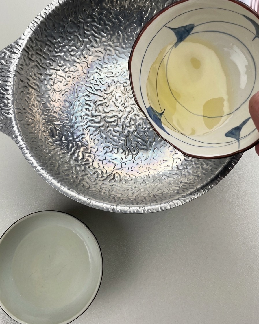 Osoba wlewająca mirin z małej ceramicznej miseczki do srebrnego garnka z fakturowaną powierzchnią, obok czeka miseczka z sake.