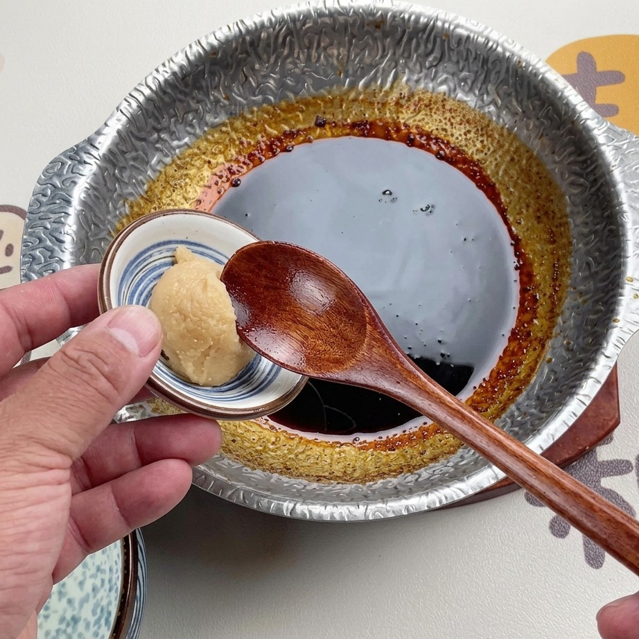 Drewniana łyżka trzymająca porcję jasnej pasty miso nad ciemnym, zredukowanym sosem w metalowym garnku.